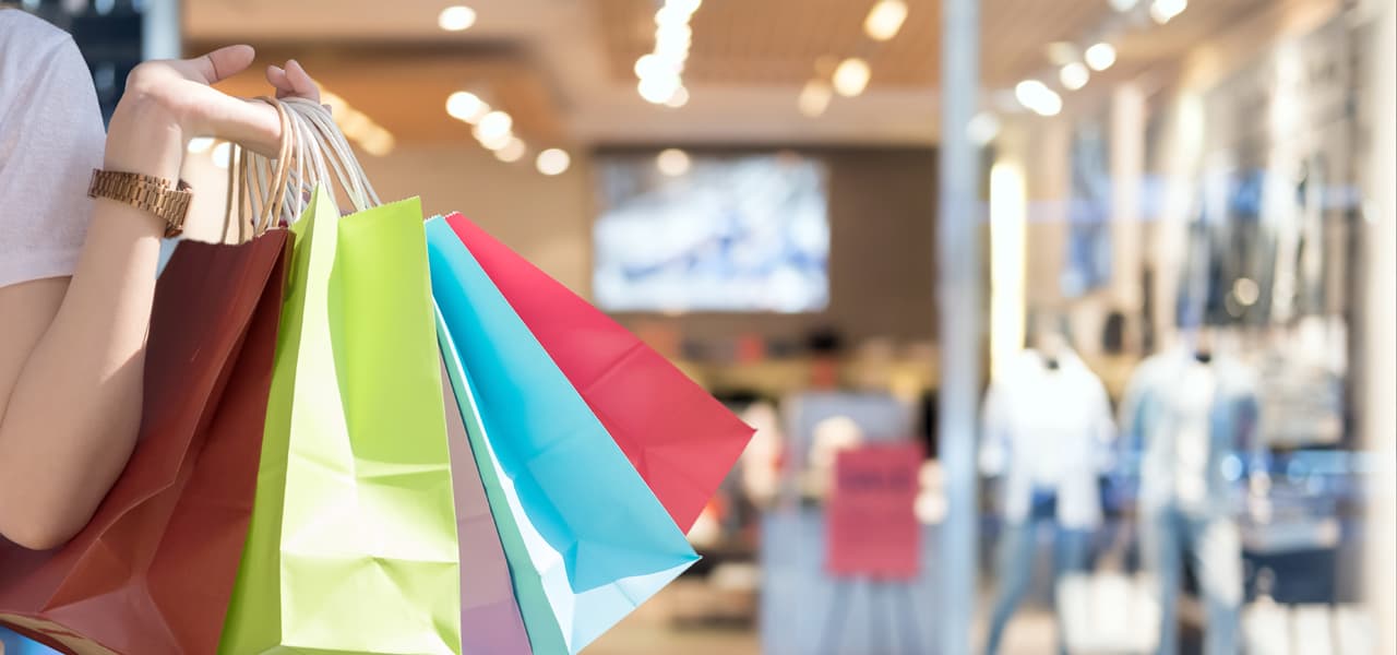 Frauenhand hält bunte Einkaufstüten.  Im Hintergrund ist ein verschwommen dargestelltes Geschäft zu sehen. | Motiv ©Natee Meepian - stock.adobe.com | © stock.adobe.com, Januar 2021