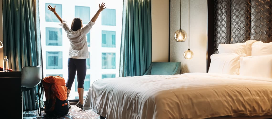 Rucksacktouristin steht am Fenster eines Hotelzimmers und reckt die Hände zum Himmel. | Motiv ©Soloviova Liudmyla - stock.adobe.com | © stock.adobe.com, September 2020